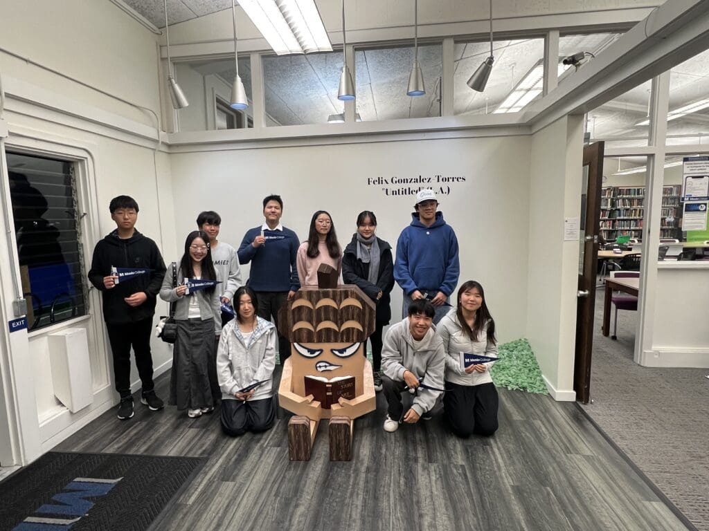 A group of students stands and kneels around the Oakie sculpture in the Bowman Library entrance area. The sculpture sits in the center, holding a book. The students hold Menlo College pennants and smile toward the camera. Bookshelves, carpeted flooring, and glass-paneled study rooms appear in the background.